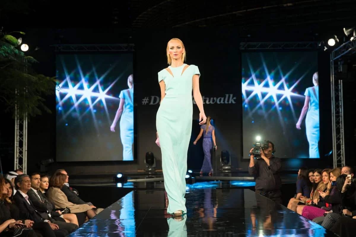 A model wearing a light blue dress walks down a glossy runway at a fashion show, with an audience seated on both sides and a cameraman capturing the moment. Two screens in the background display a reflection of the model in a similar pose.