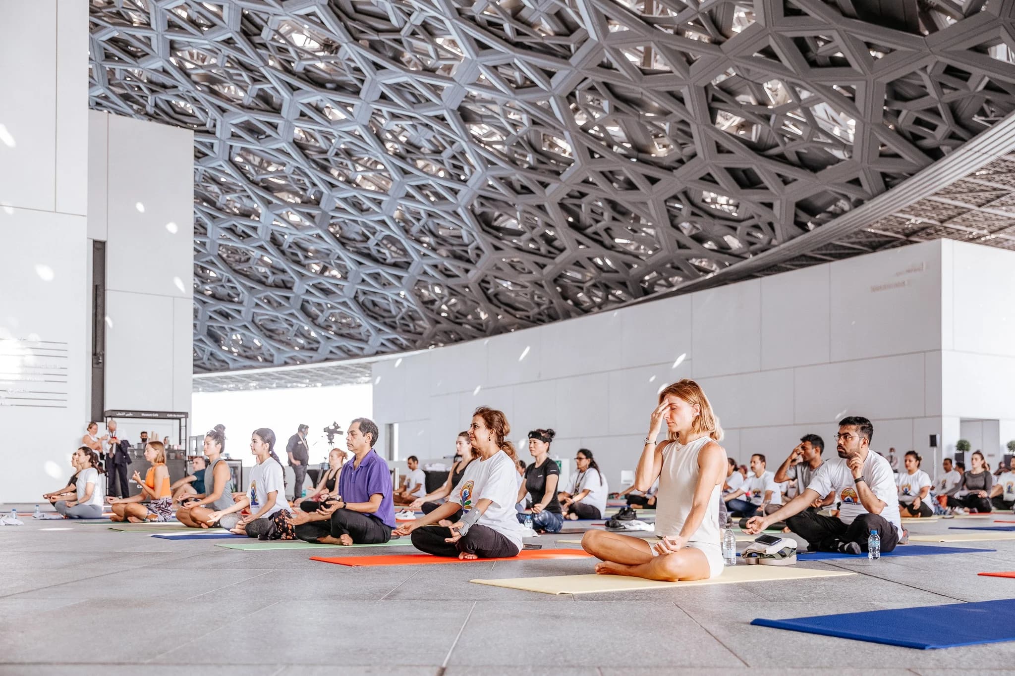 A group of people sitting cross-legged on yoga mats in a large, architecturally elaborate indoor space, practicing meditation or breathing exercises.