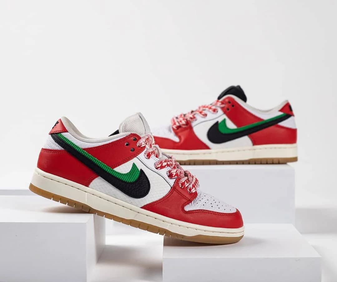 A pair of Nike sneakers displayed on white blocks. The shoes have a red, white, and green color scheme with a prominent black Nike swoosh, red and white patterned laces, and gum soles.