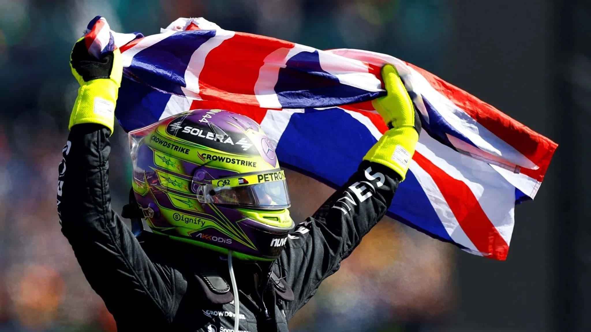 A person in racing gear with a colorful helmet and neon gloves holding a British flag overhead against a blurred background.