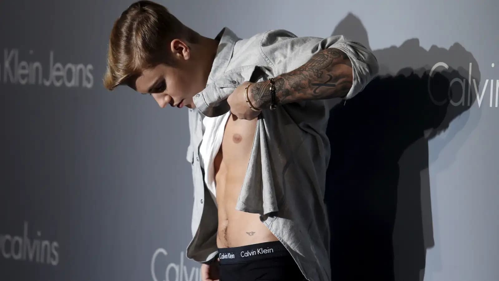 A person lifting their shirt to reveal their torso, standing in front of a Calvin Klein Jeans backdrop, with a tattoo on their arm and wearing Calvin Klein underwear.