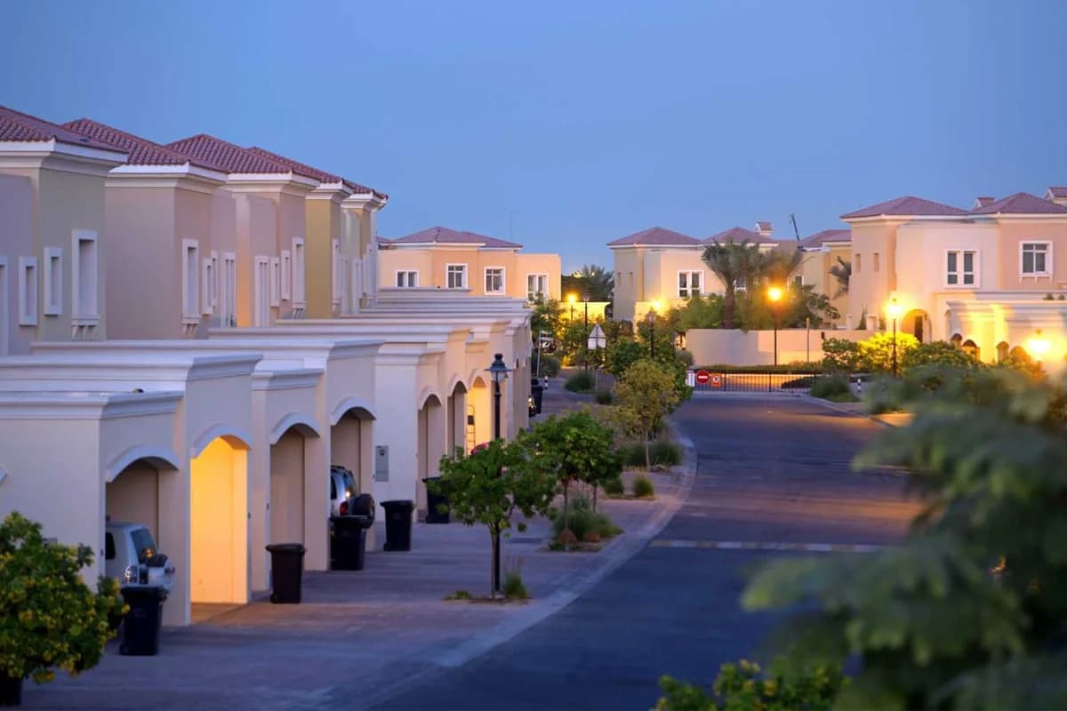 Suburban neighborhood at dusk with rows of beige stucco houses, lit streetlamps, and neatly maintained trees and shrubs along the street.