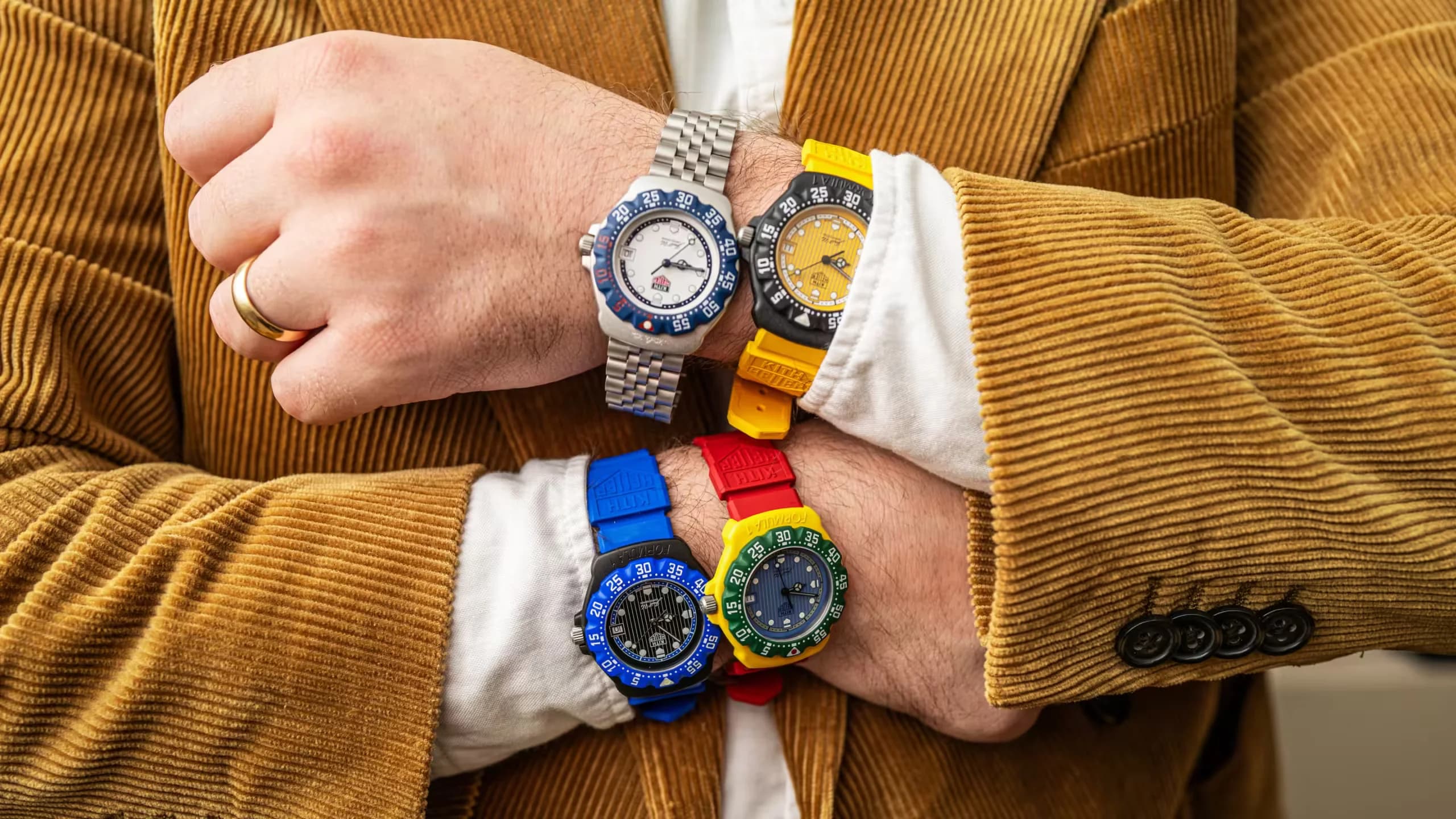 A person wearing a brown corduroy jacket shows six colorful watches on their forearms: one with a metal band and the others with bright blue, red, yellow, green, and black straps. The person also wears a gold ring on their finger.