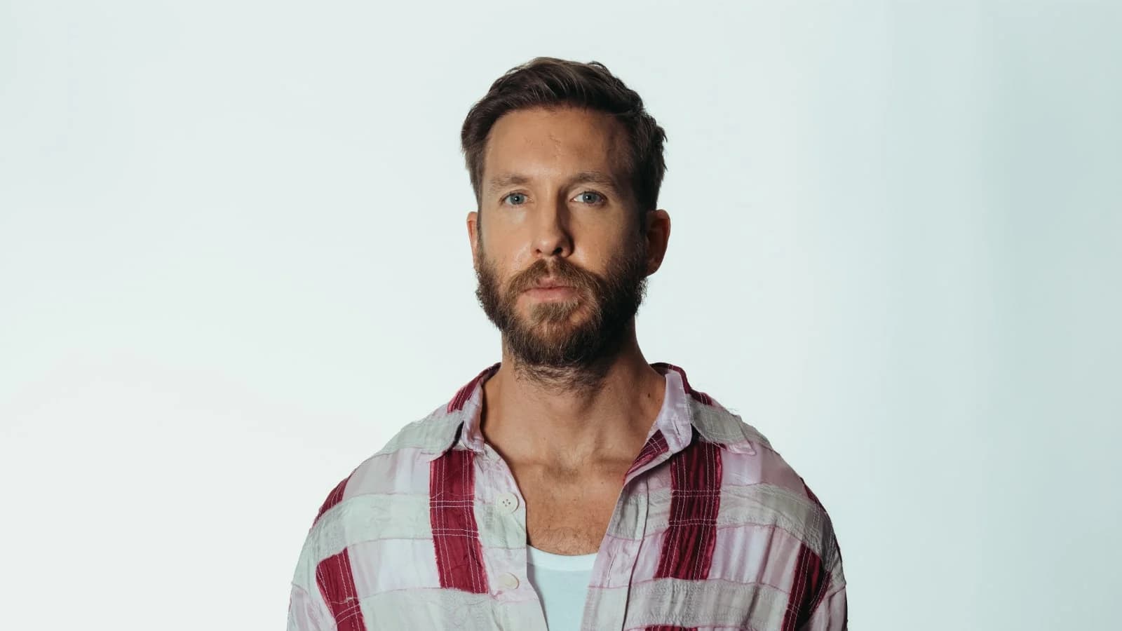 A man with a beard and short hair, wearing a plaid shirt and a white undershirt, standing against a plain white background.