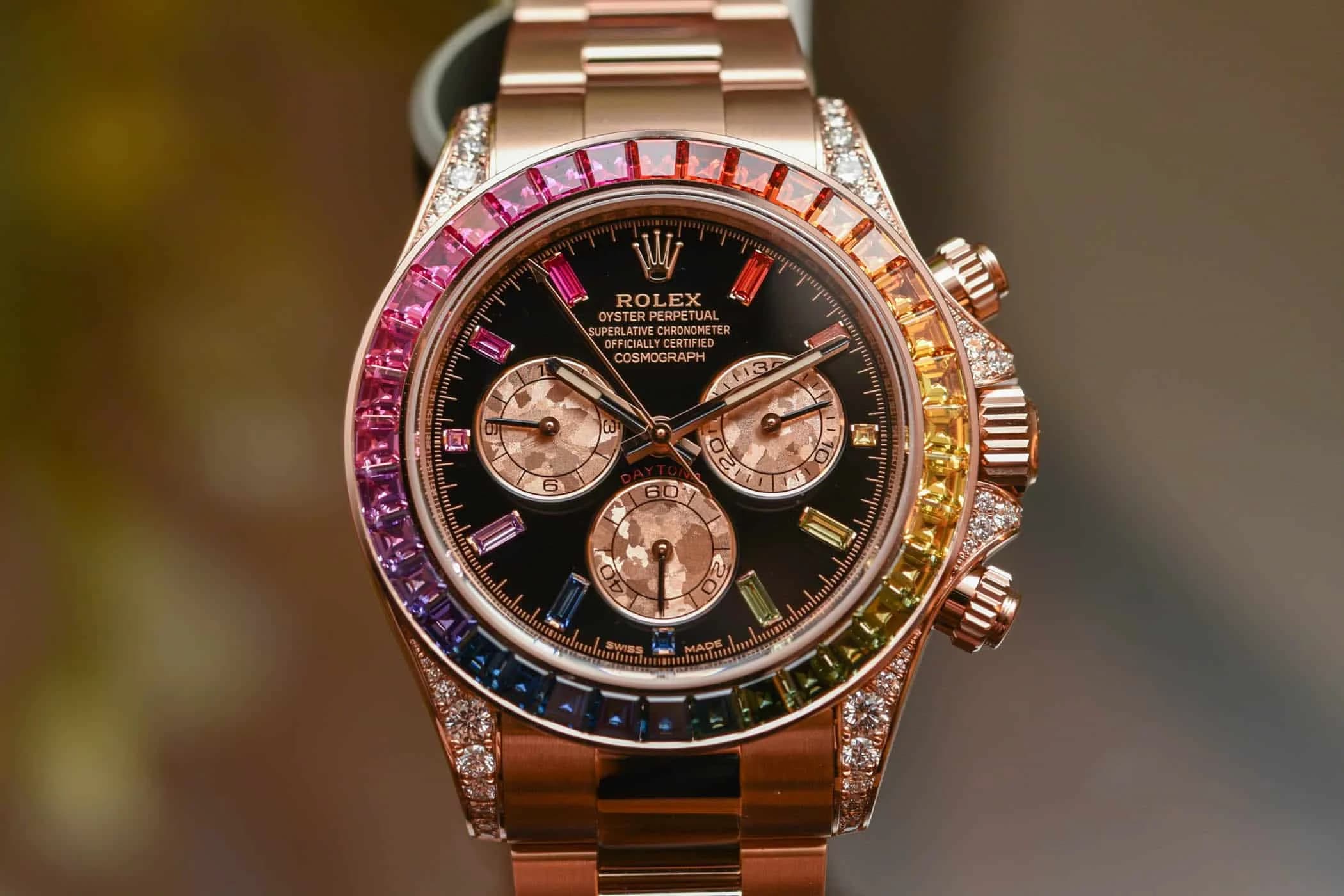 A luxurious wristwatch with a rose gold strap and a bezel adorned with multicolored gemstones. The black watch face features three sub-dials and diamond markers, with additional diamond embellishments on the case.