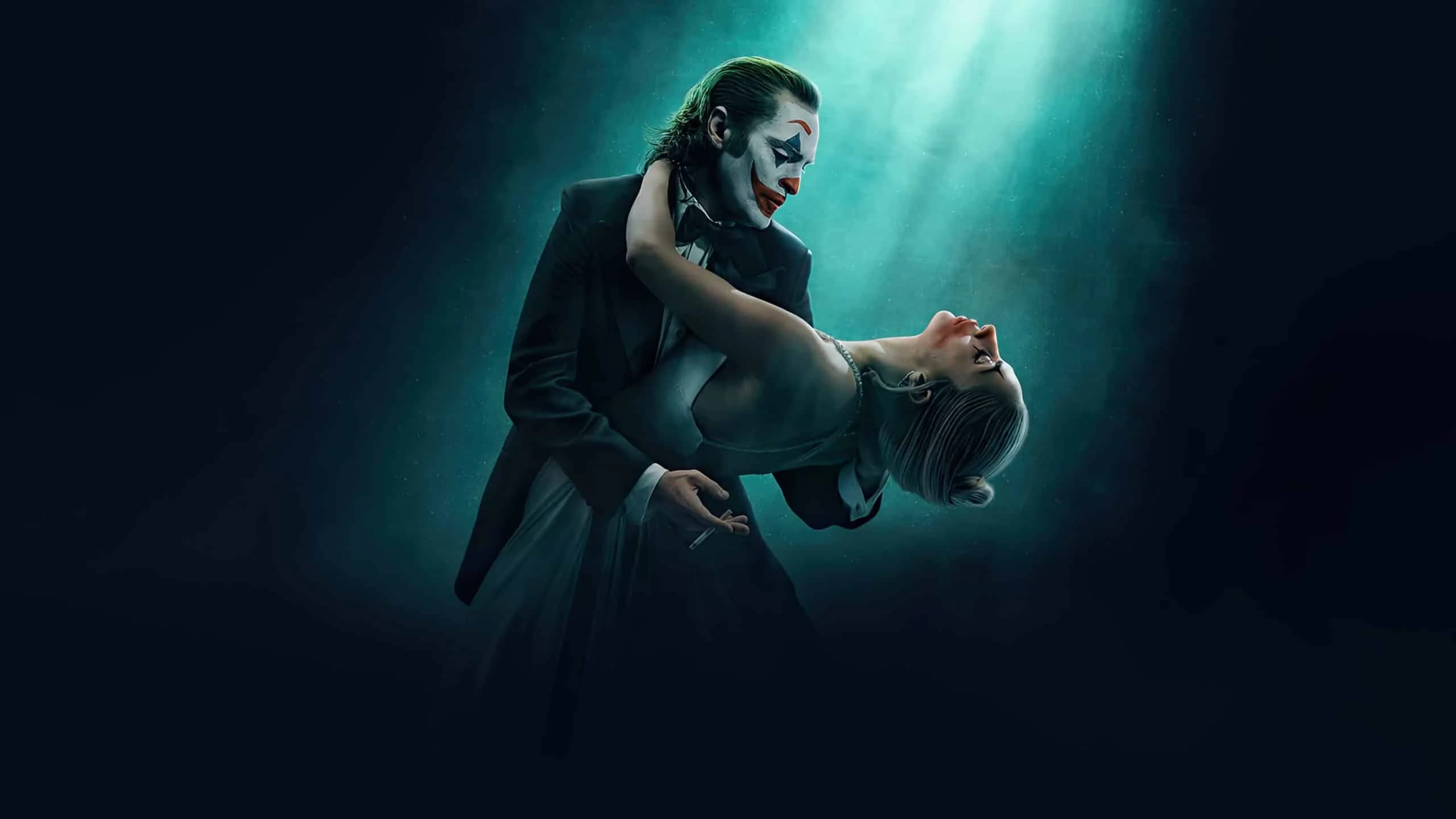 A person dressed in a dark suit with clown makeup holds another person wearing a light-colored dress and pearl necklace in a dance pose under a dramatic spotlight.