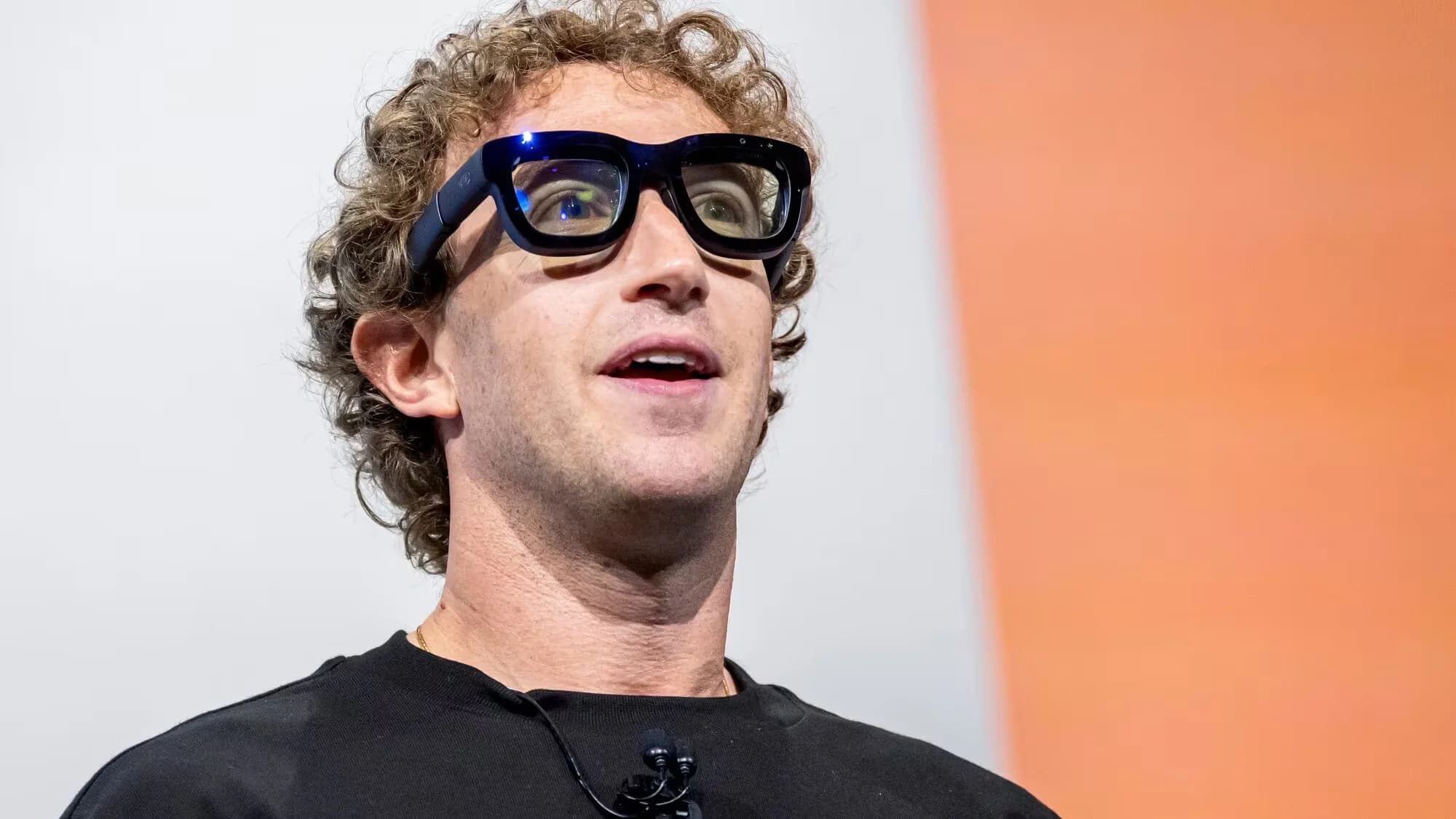 A person wearing augmented reality glasses and a black shirt, standing with an engaging expression against a backdrop with orange and white panels.