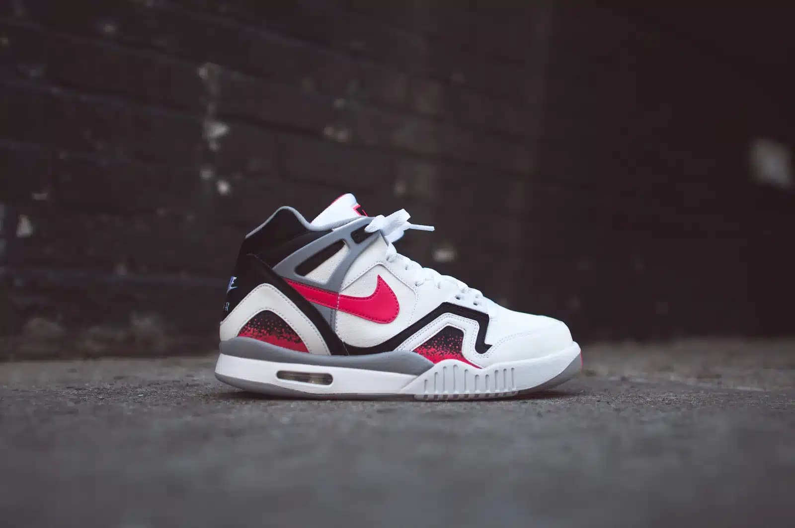 A side view of a white and black sneaker with red accents, featuring a prominent red swoosh logo, resting on a concrete surface against a dark, blurred background.