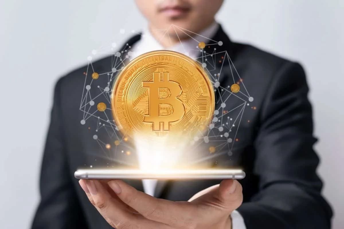 A person in a suit is holding a smartphone, with a large digital representation of a Bitcoin coin hovering above the screen, surrounded by abstract network connections and glowing light effects.