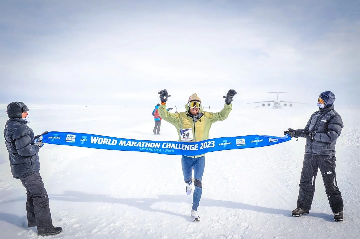 A runner celebrating as they cross the finish line of the World Marathon Challenge 2023 in Antarctica, with two people in winter gear holding the blue race banner in snowy conditions.