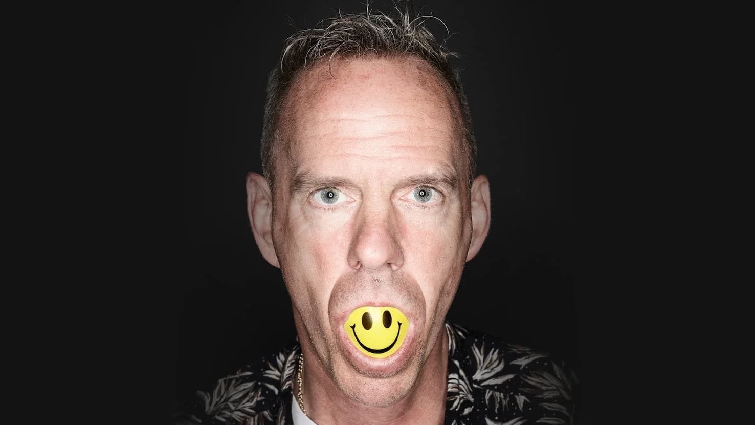 A man with short hair is looking directly at the camera with a yellow smiley face ball in his mouth against a dark background.