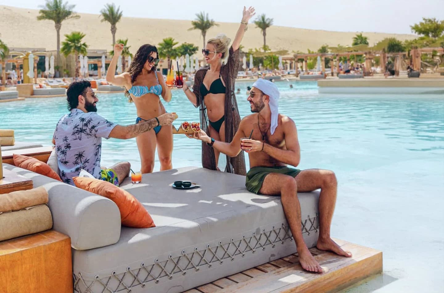 A lively poolside scene featuring four friends enjoying drinks and snacks. Two women in swimsuits and sunglasses are playfully interacting, while two men sit on a lounge, one gesturing with food and the other holding a drink. The pool is surrounded by palm trees and sunny skies, creating a relaxed vacation vibe.