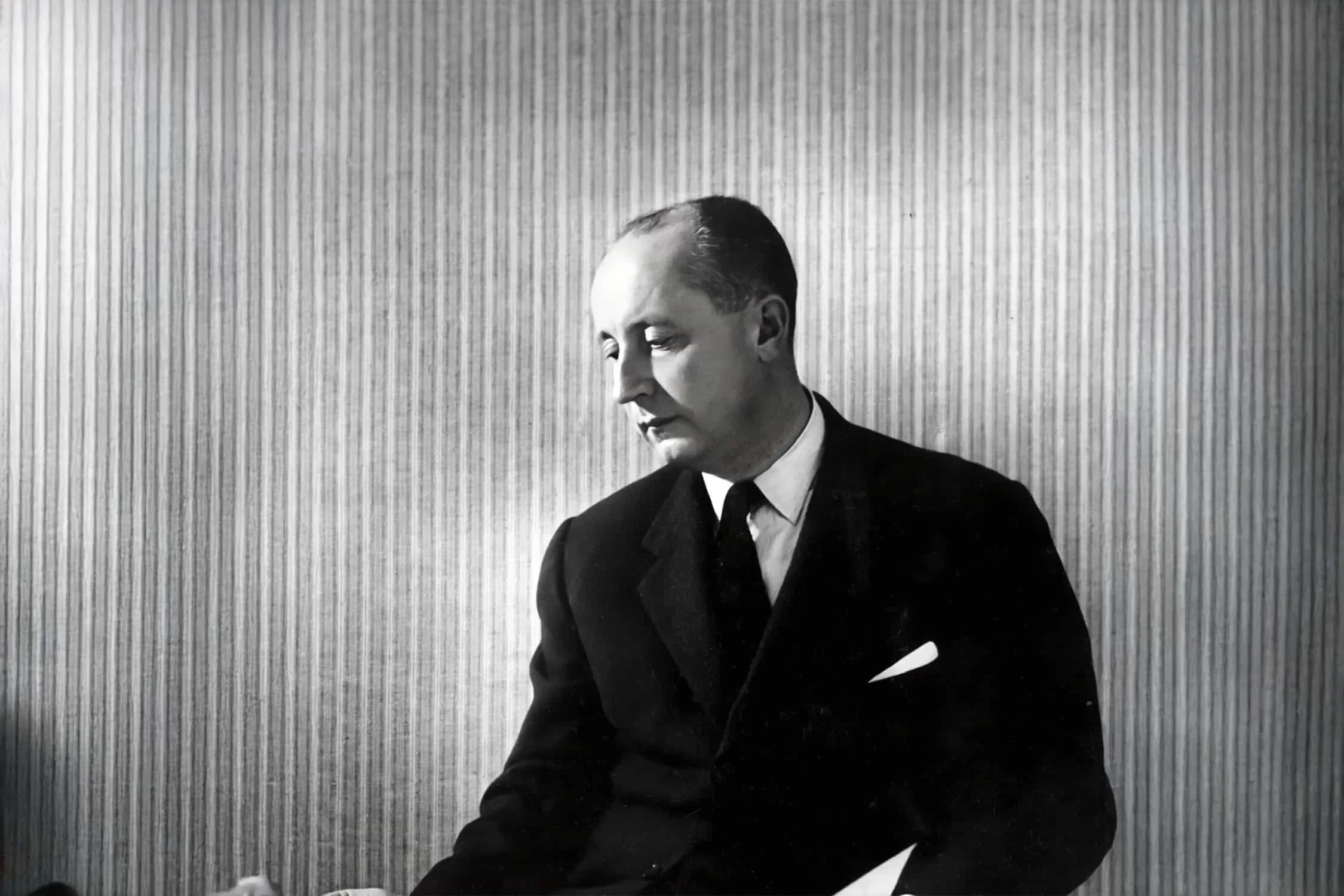 A black and white portrait of a man in a suit, looking downward against a vertically striped background.