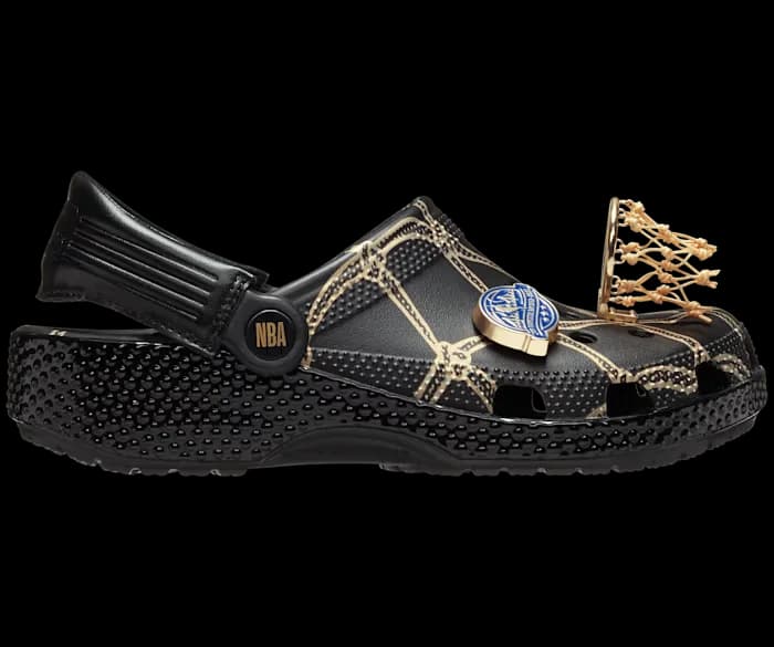 A black Crocs shoe with unique basketball-themed decorations, including a textured upper resembling a basketball court, an NBA logo on the side, and a miniature basketball hoop charm near the toe area.