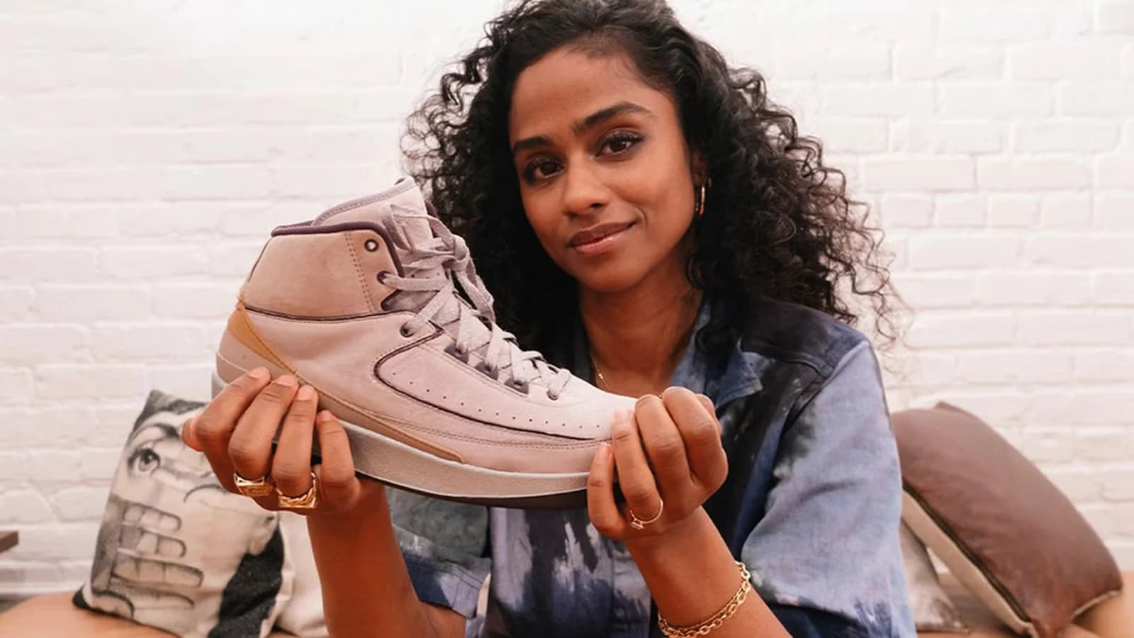 A person with curly hair holding a beige high-top sneaker, standing against a white brick wall.
