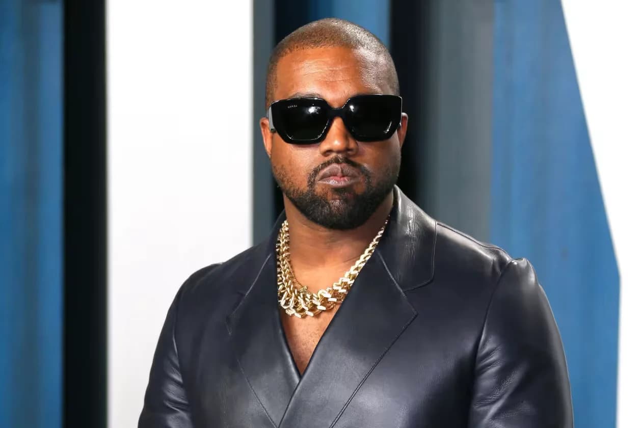 A man wearing dark sunglasses and a black leather jacket with a gold chain necklace in front of a blue and white background.
