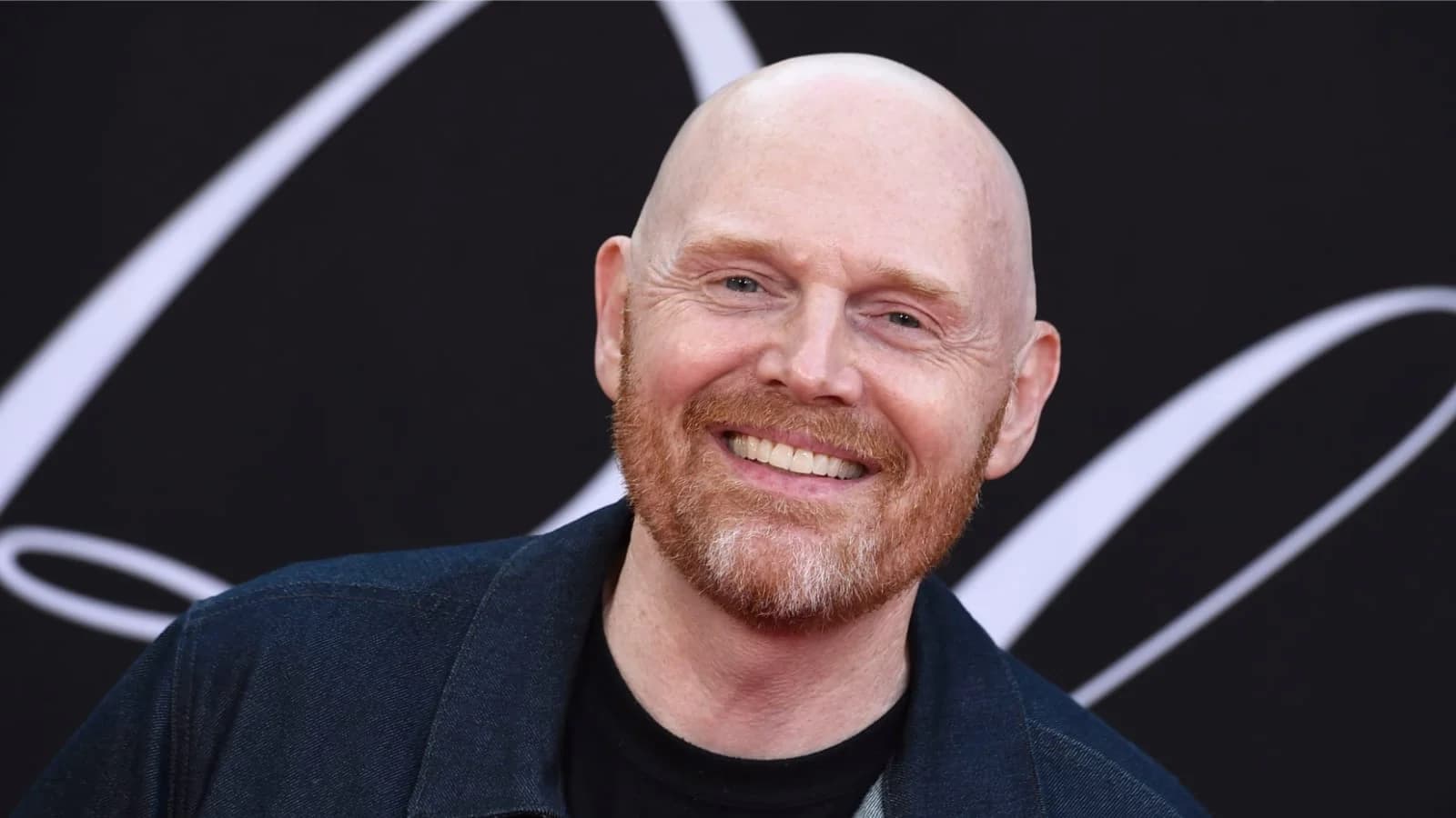 A bald man with a red beard smiling in front of a black background with white lines.