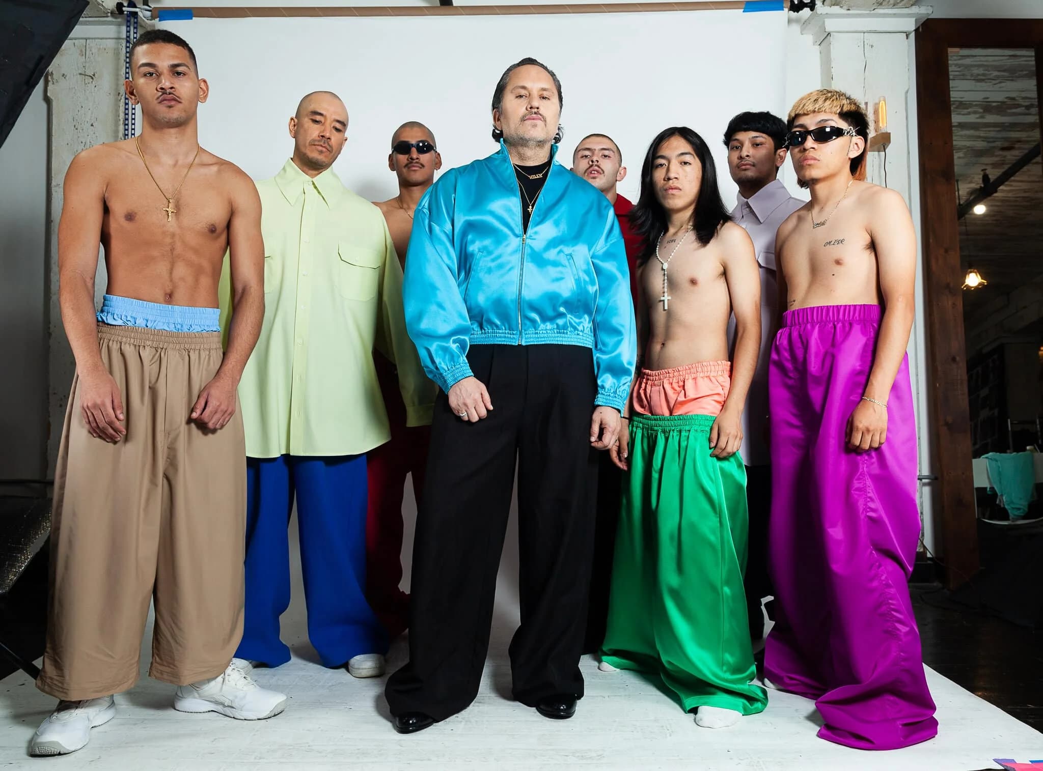 A group of men posing indoors, wearing brightly colored, oversized pants; some are topless, and others wear loose jackets and shirts. The background shows a plain wall and studio lighting equipment.