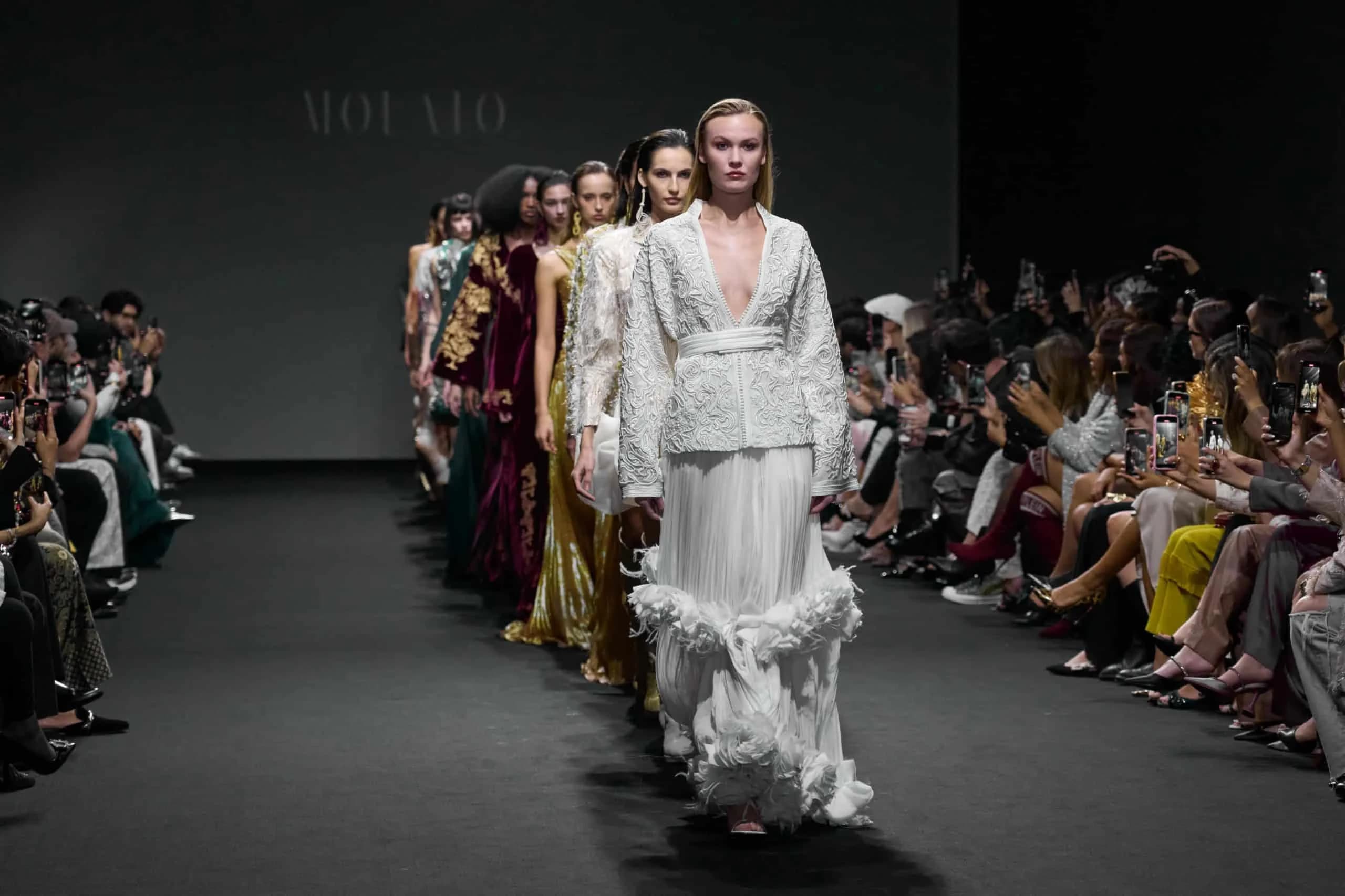 A group of models walking down the runway in elegant gowns during a fashion show, with a seated audience capturing photos on their phones on both sides.