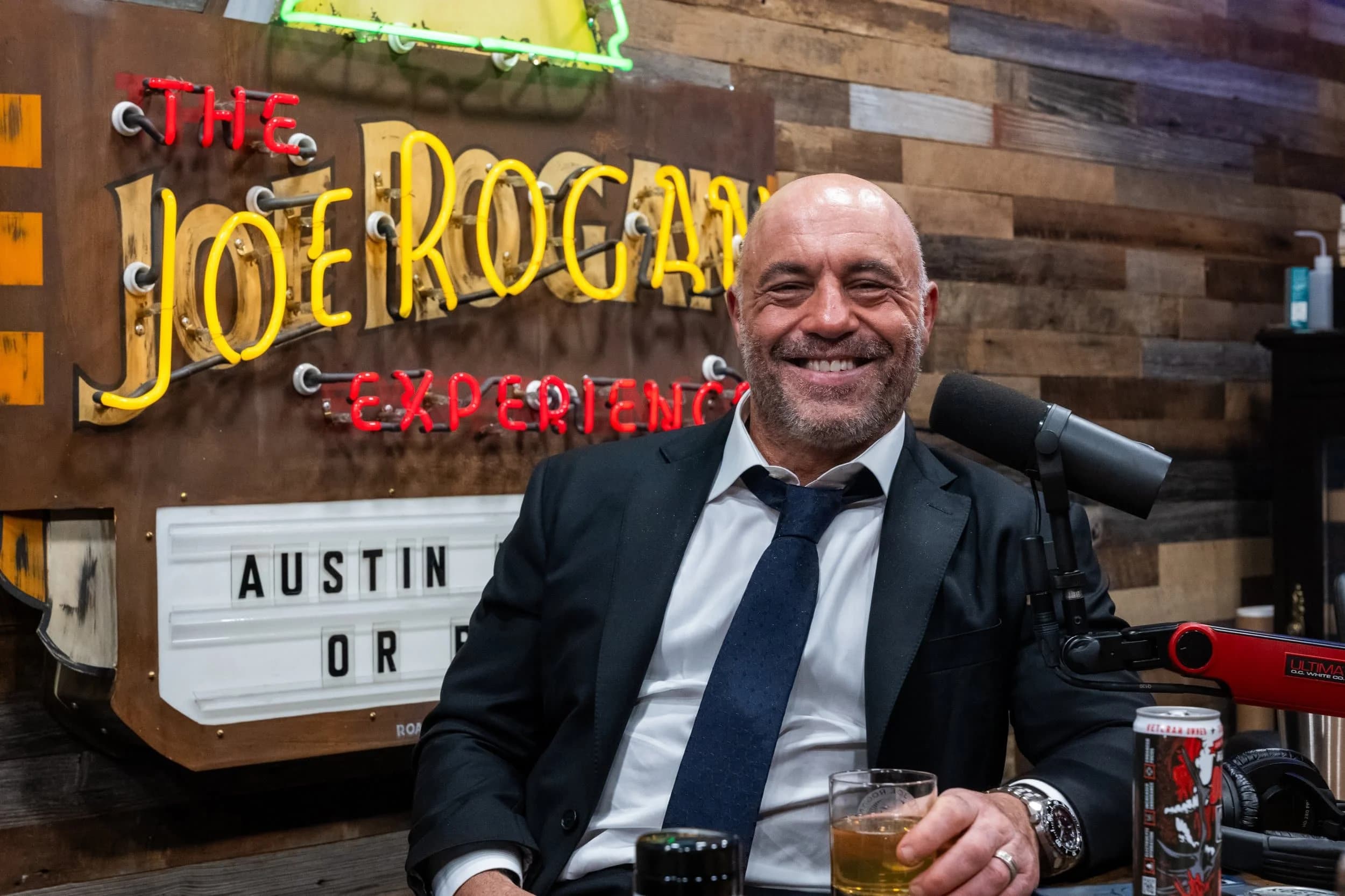 A man in a suit and tie sits smiling at a podcast desk, holding a glass of drink with a microphone in front of him and a neon sign reading "The Joe Rogan Experience" in the background.