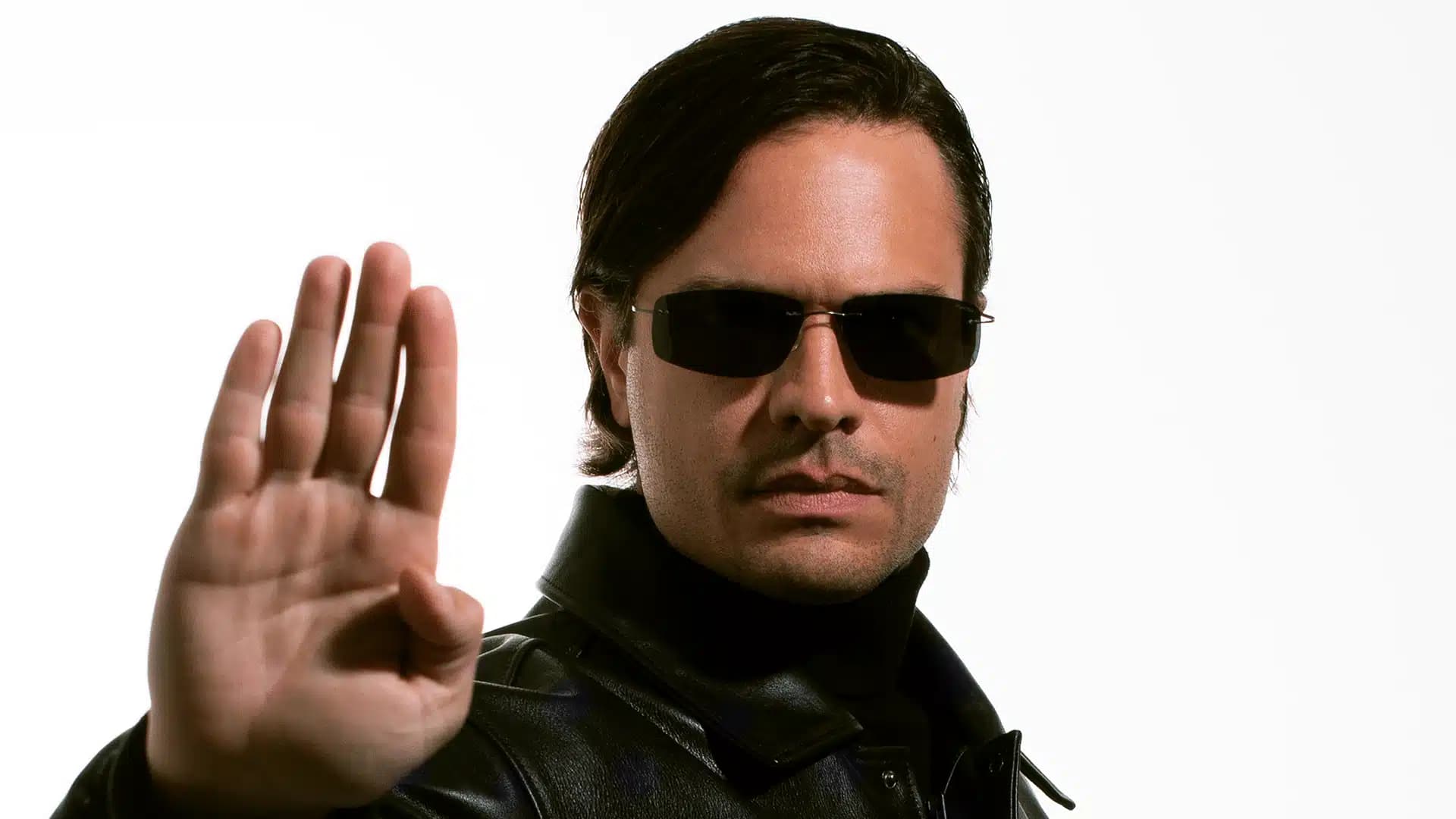 A man wearing sunglasses and a black leather jacket stands against a white background, holding his hand up in a stop gesture.
