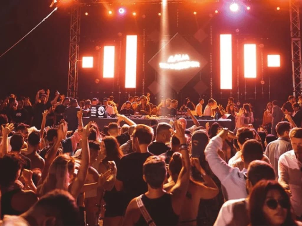 A lively crowd dancing at a music event, with vibrant stage lights and a DJ booth in the background, highlighting an energetic atmosphere filled with raised hands and diverse attendees.