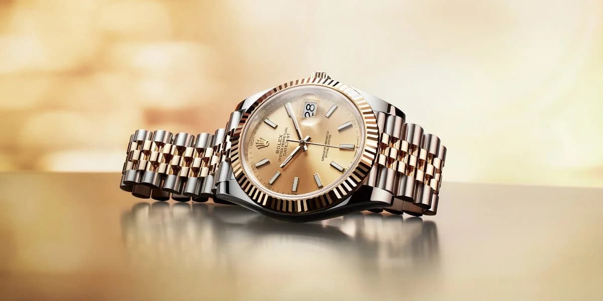 A luxury wristwatch with a champagne-colored dial, featuring a fluted bezel, a date window, and a two-tone metal bracelet combining gold and stainless steel, displayed on a reflective surface with a soft, blurred background.