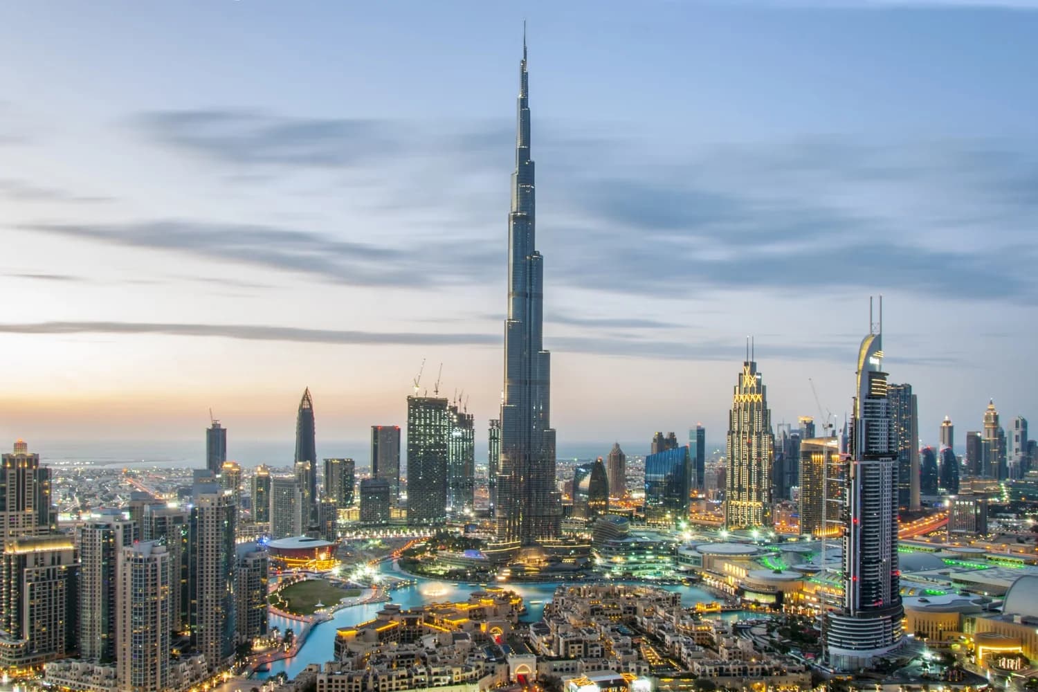 A panoramic view of Dubai's skyline at dusk, featuring the Burj Khalifa prominently in the center, surrounded by various modern skyscrapers and illuminated buildings, with waterways and greenery below.