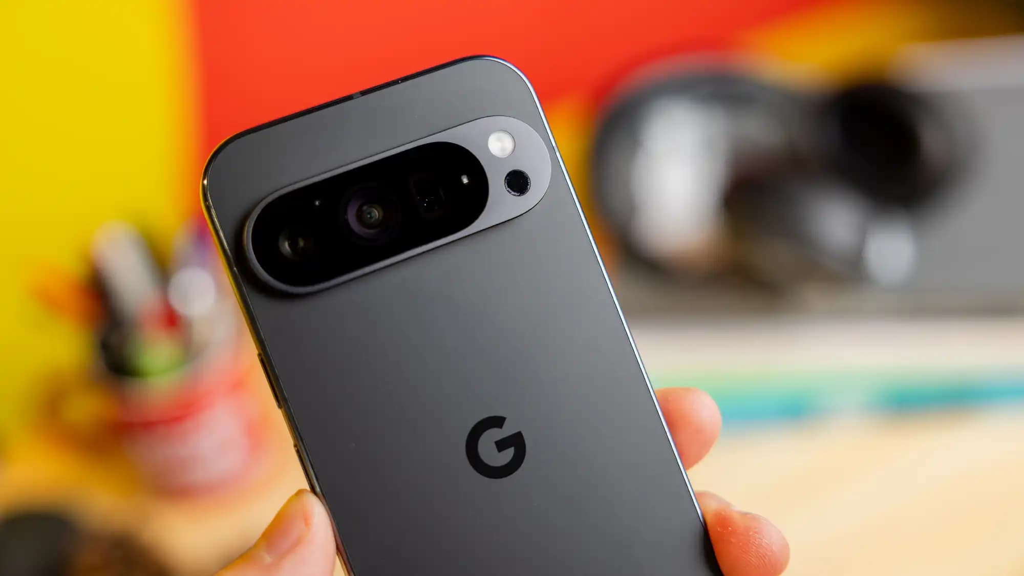 Close-up of the back of a smartphone held in a hand, featuring a prominent camera module with multiple lenses and a flash, against a blurred background of vibrant colors and various office supplies.