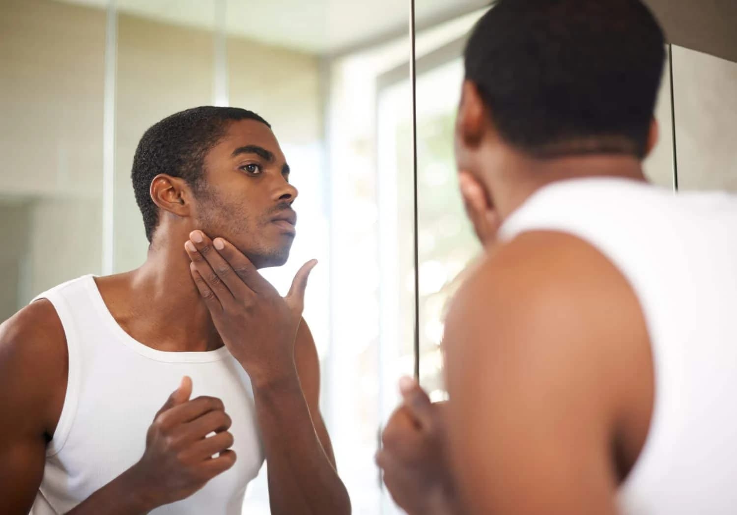 A man in a white tank top stands in front of a mirror, inspecting his face and grooming his facial hair.
