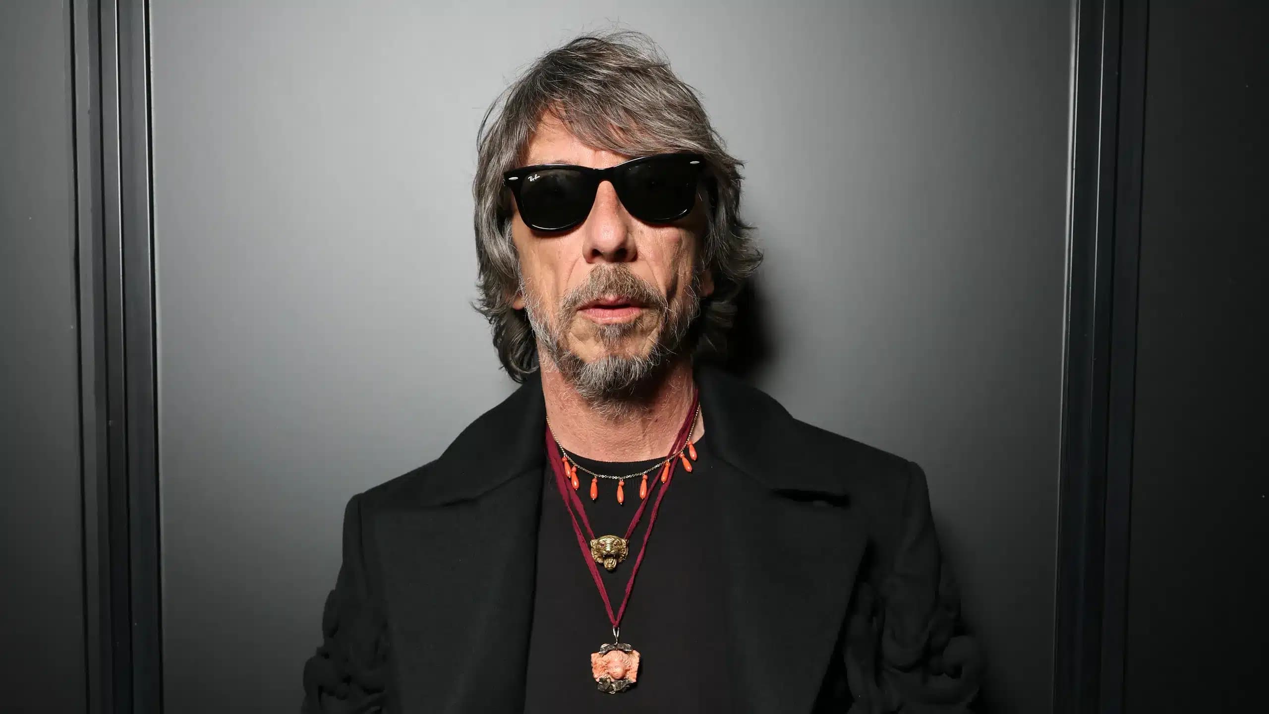 A man with shaggy gray hair and sunglasses stands against a dark background. He wears a black coat and multiple necklaces with various pendants.