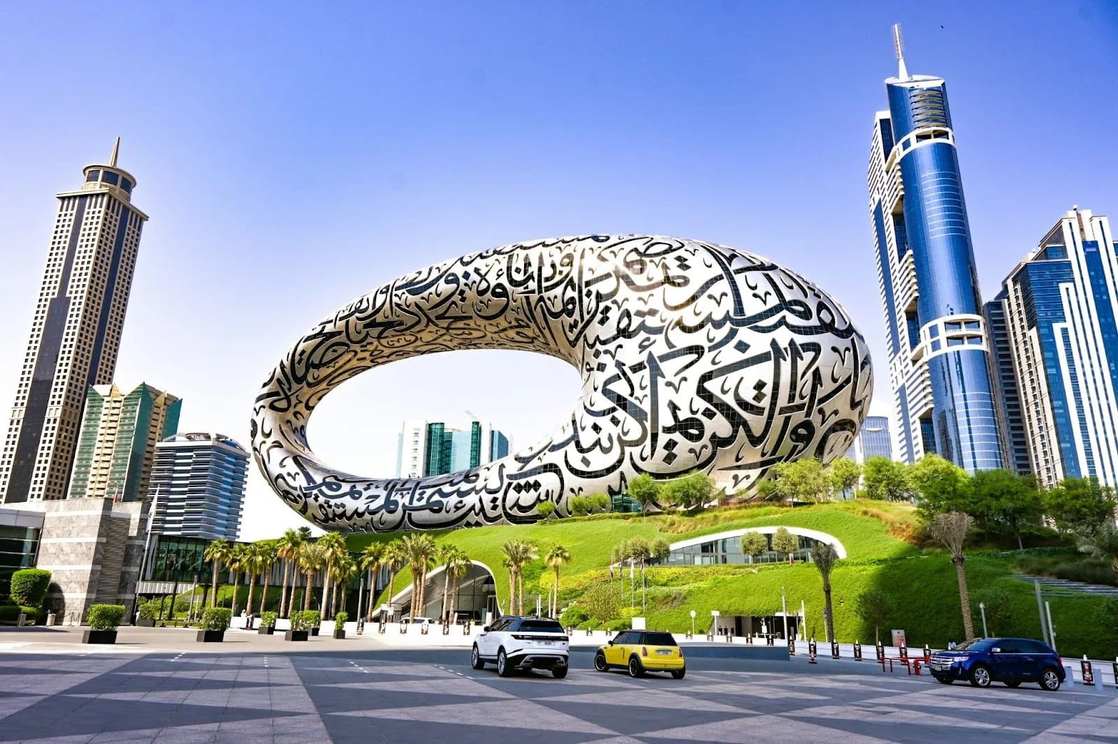A futuristic building with a large, oval structure covered in Arabic calligraphy, set against a backdrop of modern high-rise buildings and palm trees, with cars parked in the foreground on a paved area.