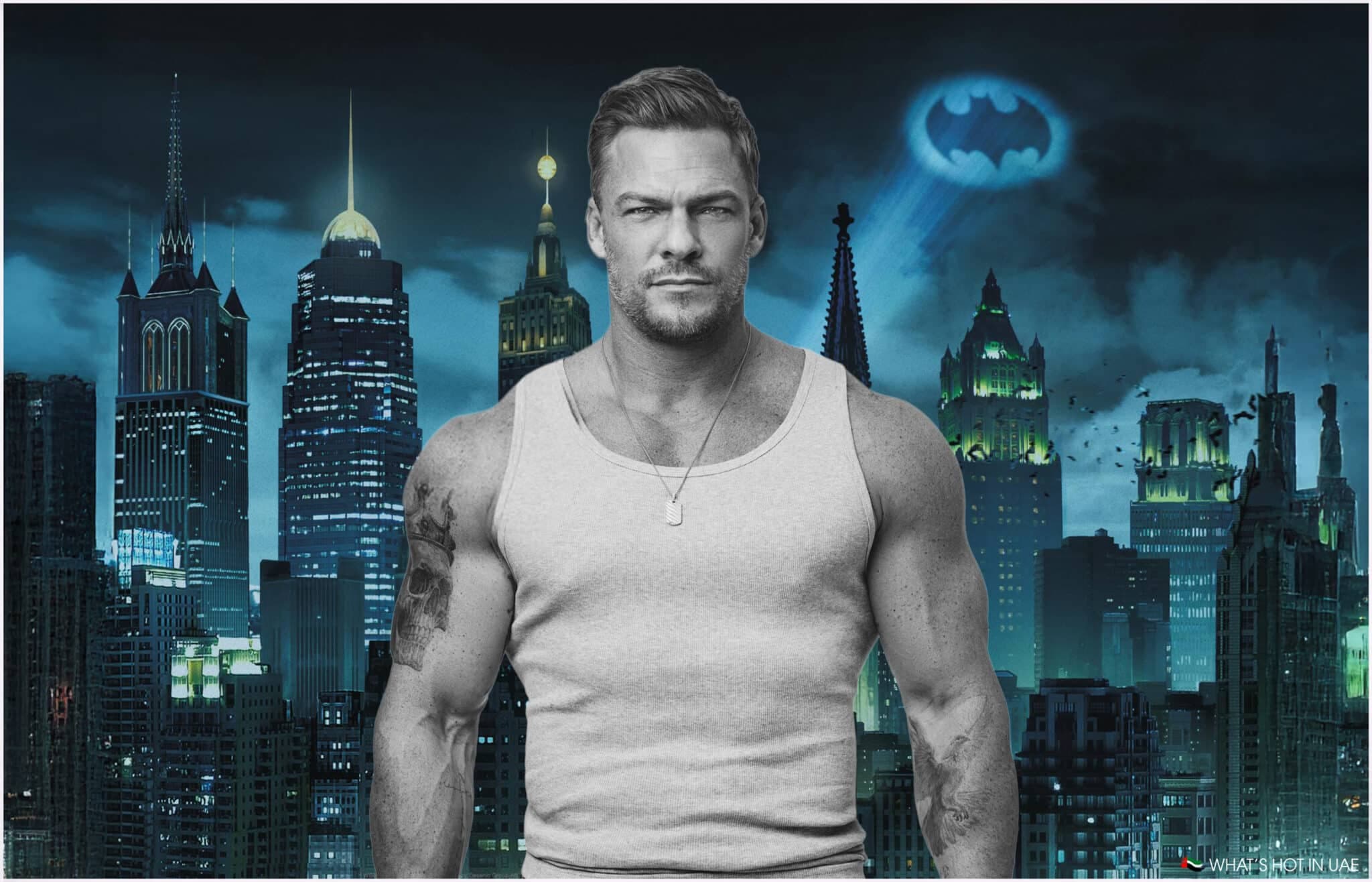 A muscular Alan Ritchson wearing a gray tank top stands in front of a city skyline at night, featuring skyscrapers and the Bat-Signal projected into the sky.