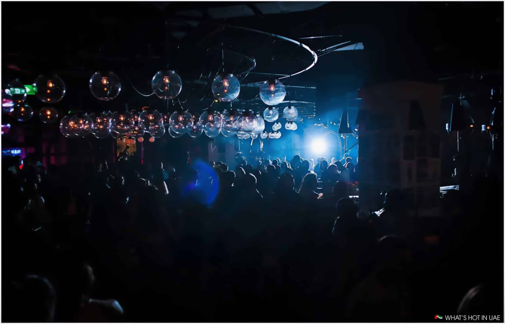 A lively nightclub scene with a crowded dance floor, dim lighting, and numerous hanging spherical lights, set against a backdrop of blue and purple hues.