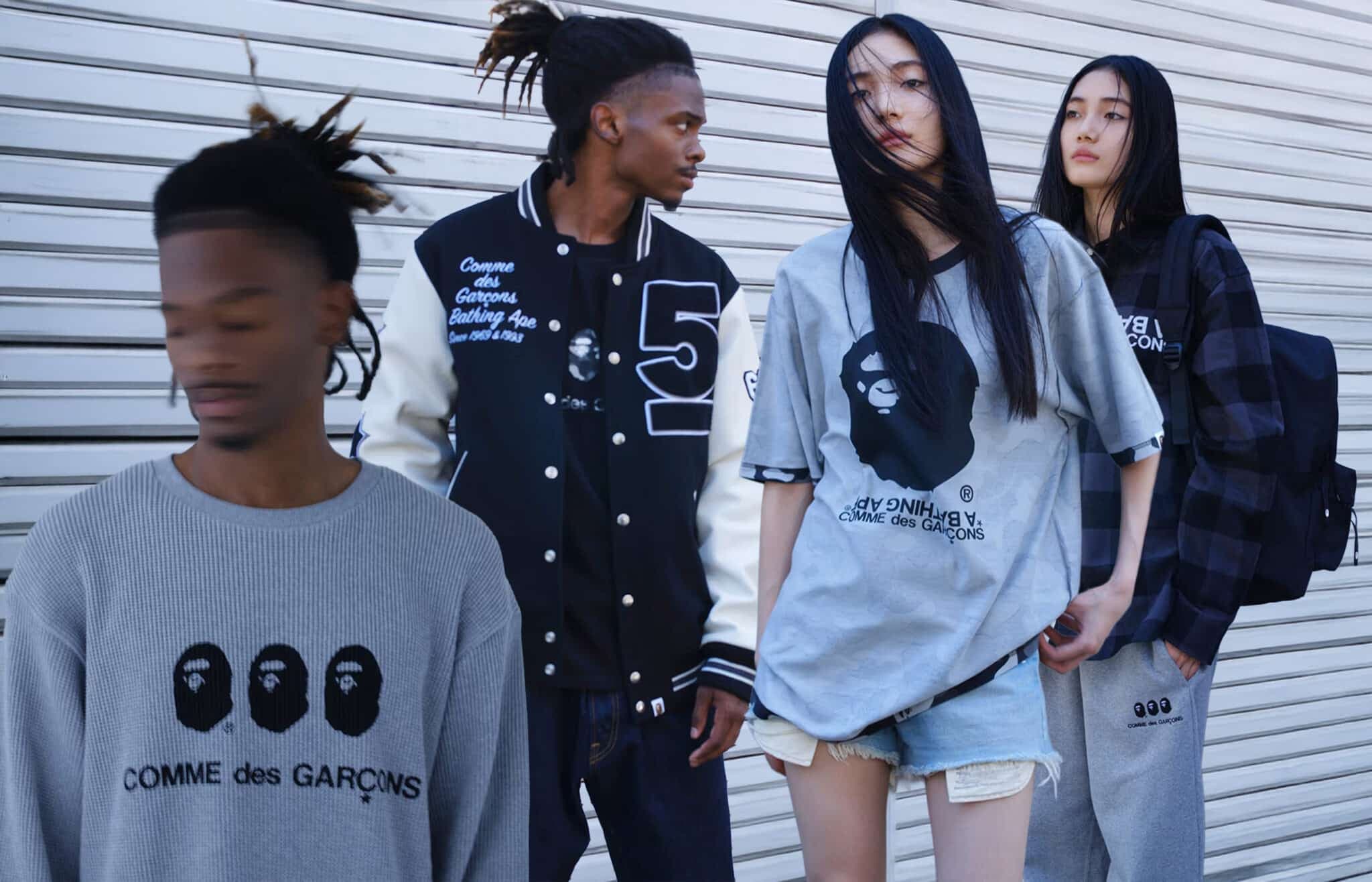 Four people are standing in front of a metal shutter, wearing BAPE clothes featuring "Comme des Garçons" and stylized face designs. The person in the foreground has blurred features and a grey sweater, while others wear a varsity jacket, oversized t-shirts, and casual pants.