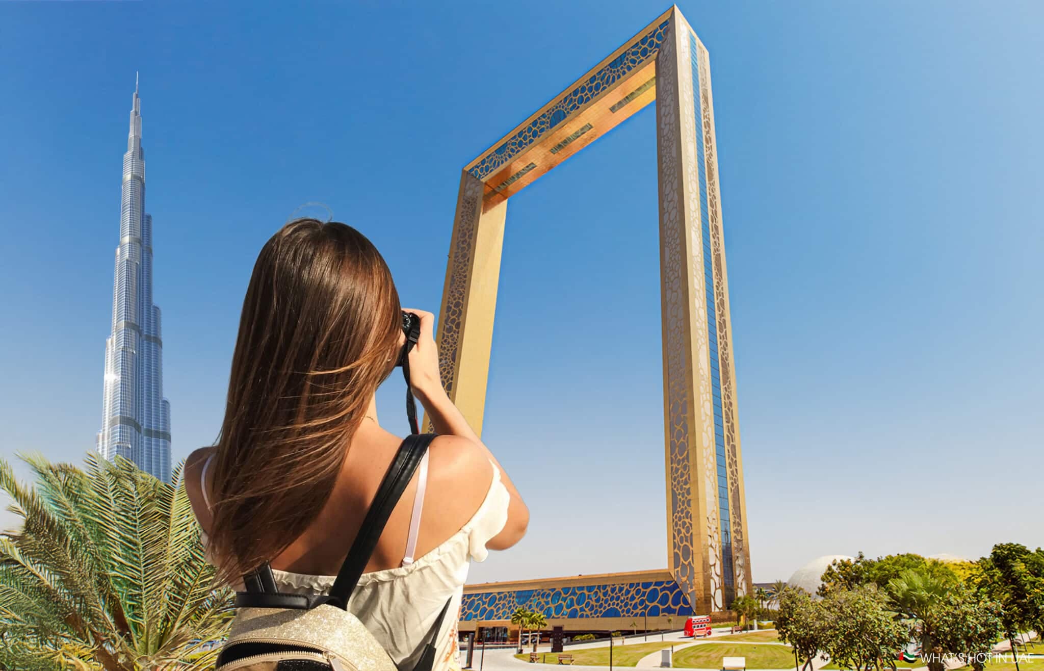 A woman with long brown hair is taking a photo of the Dubai Frame with a camera. The Burj Khalifa is visible in the background against a clear blue sky.