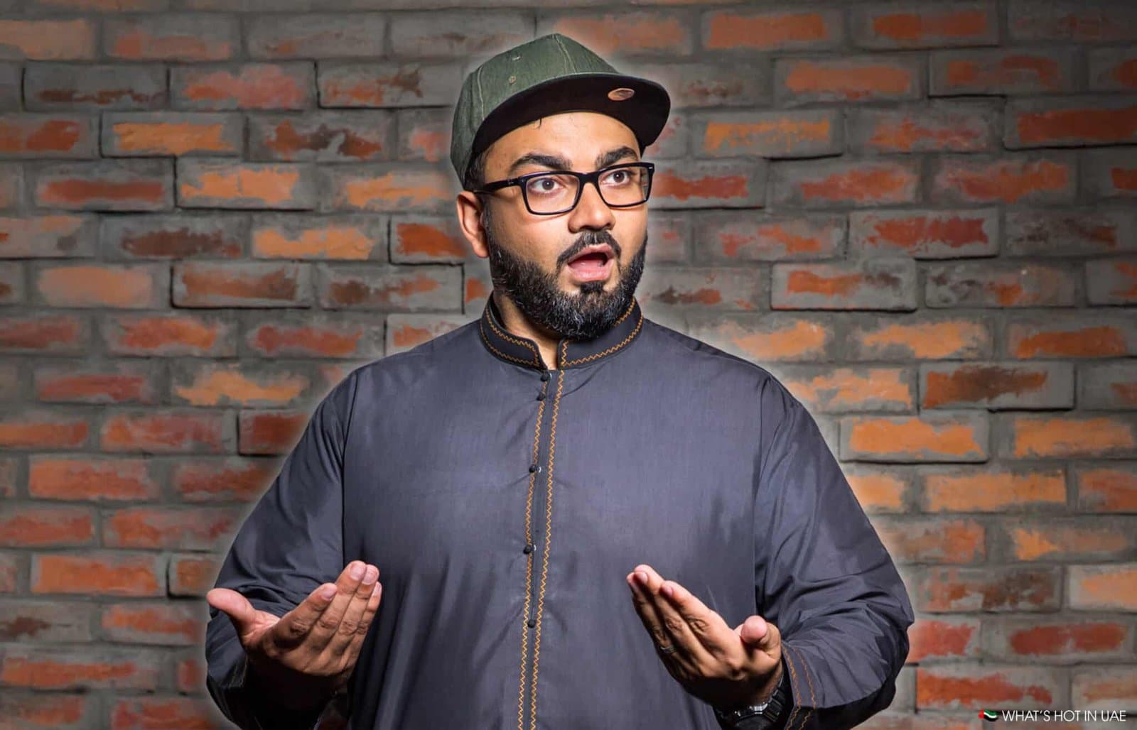 A man wearing a dark traditional outfit and a cap, with an expressive facial expression, stands in front of a brick wall background.