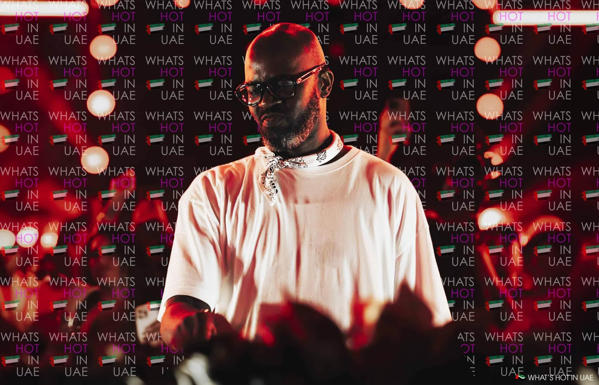 A man in a white shirt and bandana stands in front of a backdrop with 'What's Hot in UAE' text and UAE flags, surrounded by circular lights.
