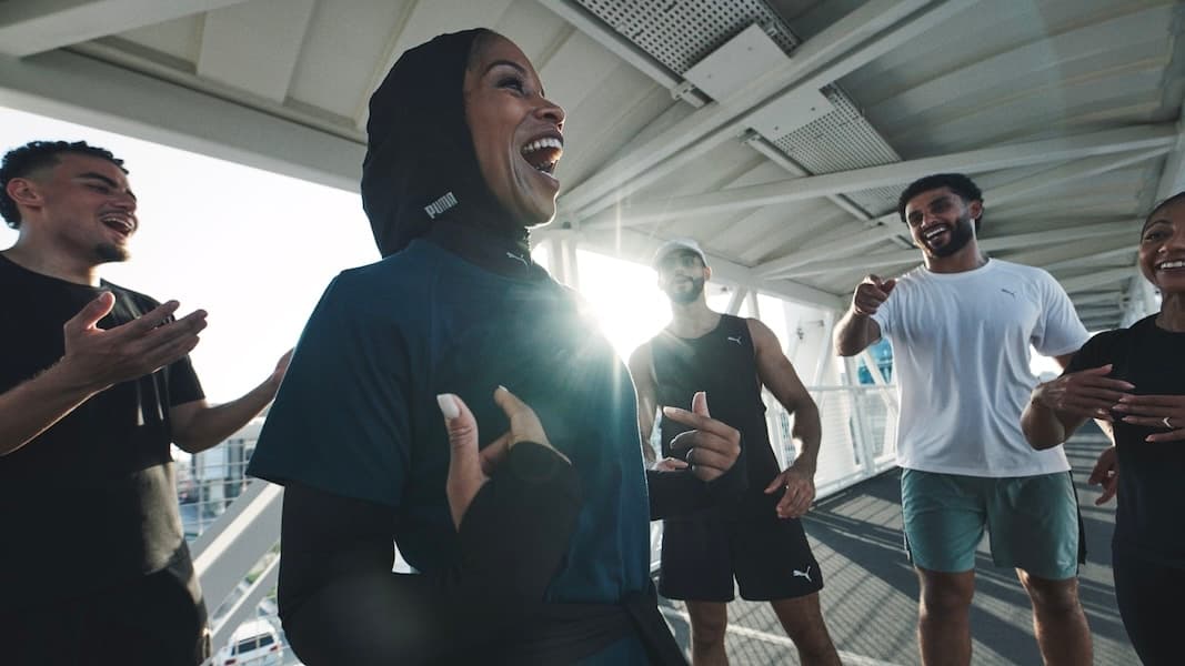 A group of people in athletic wear are laughing and engaging energetically with each other on a covered outdoor walkway in the sunlight.