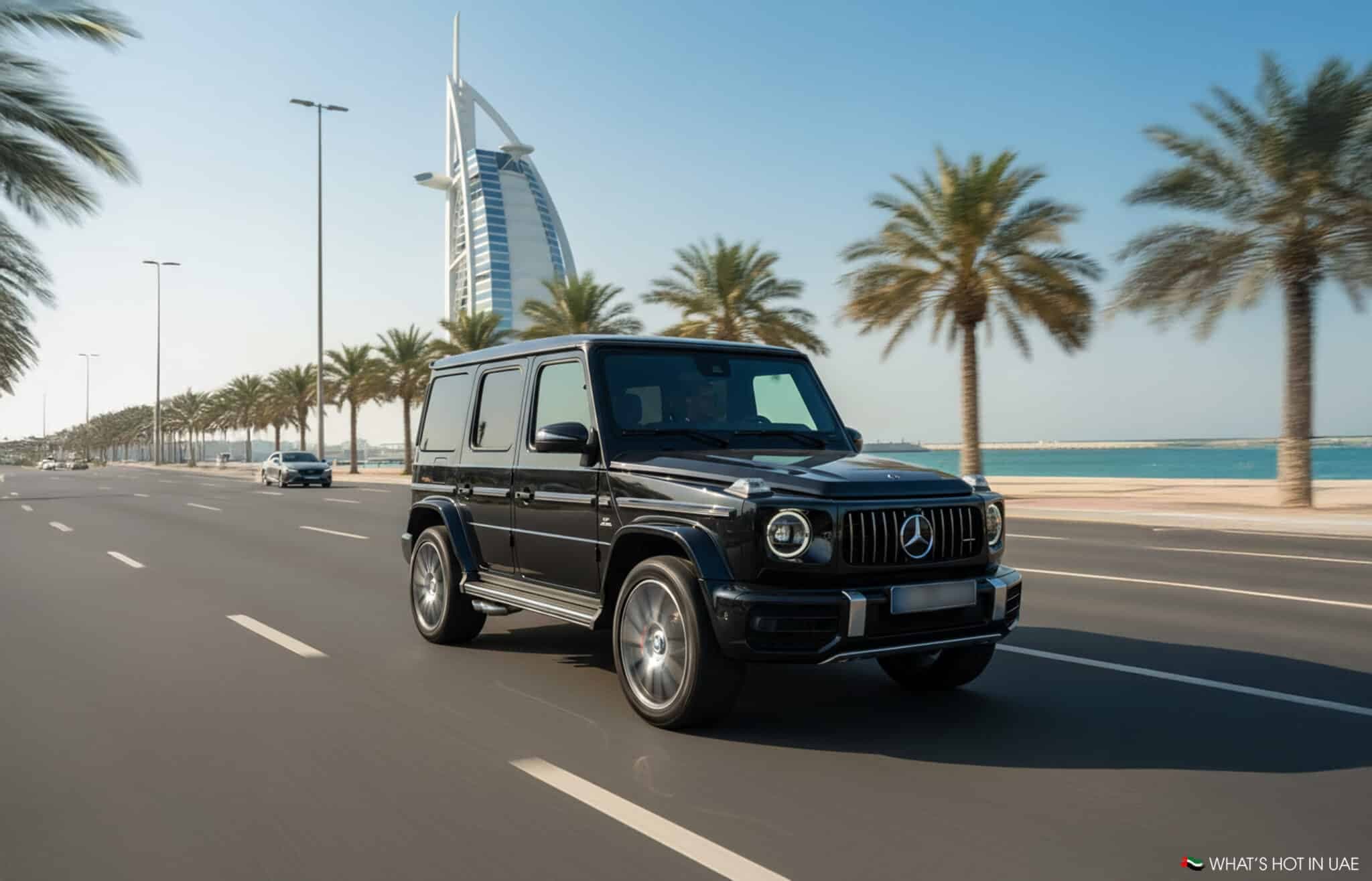 A black Mercedes-Benz G-Class SUV driving on a wide road lined with palm trees, with the Burj Al Arab in the background against a clear blue sky in Dubai, UAE.