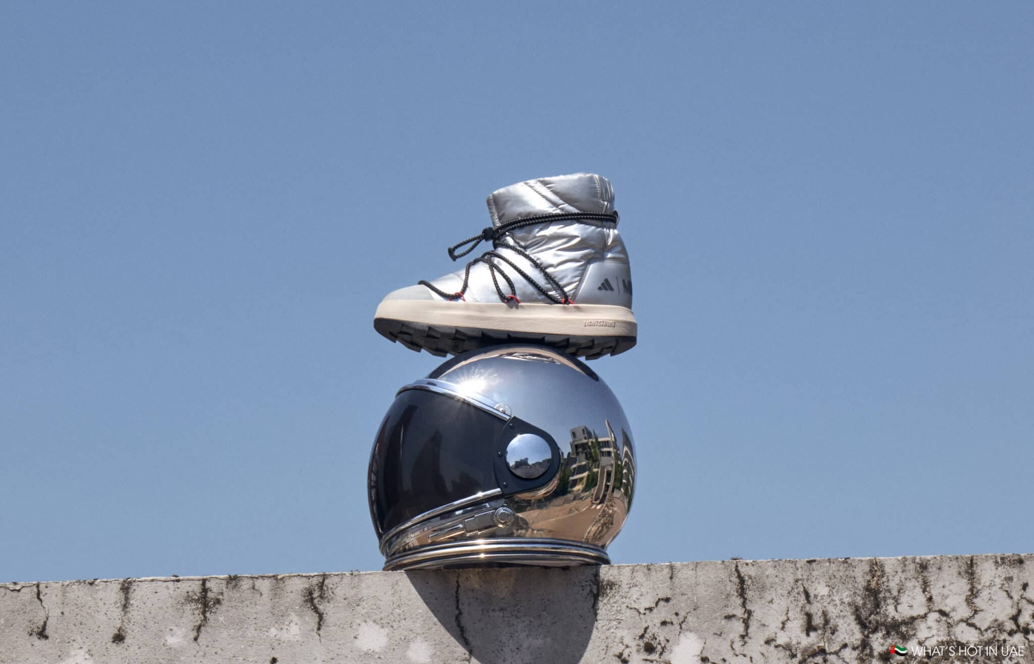A silver boot sits atop a reflective, chrome astronaut helmet on a concrete ledge against a clear blue sky.