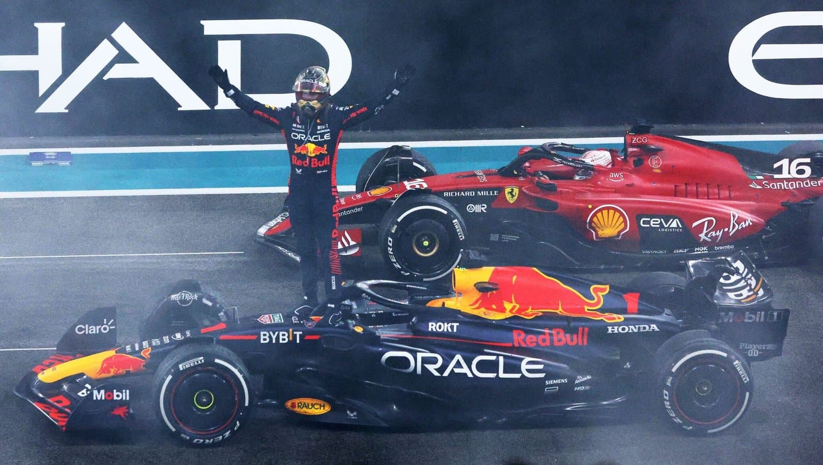 A Formula 1 driver in a Red Bull racing suit stands beside a Red Bull race car during a pause in an event, with a Ferrari race car in the background, and track signage visible above.