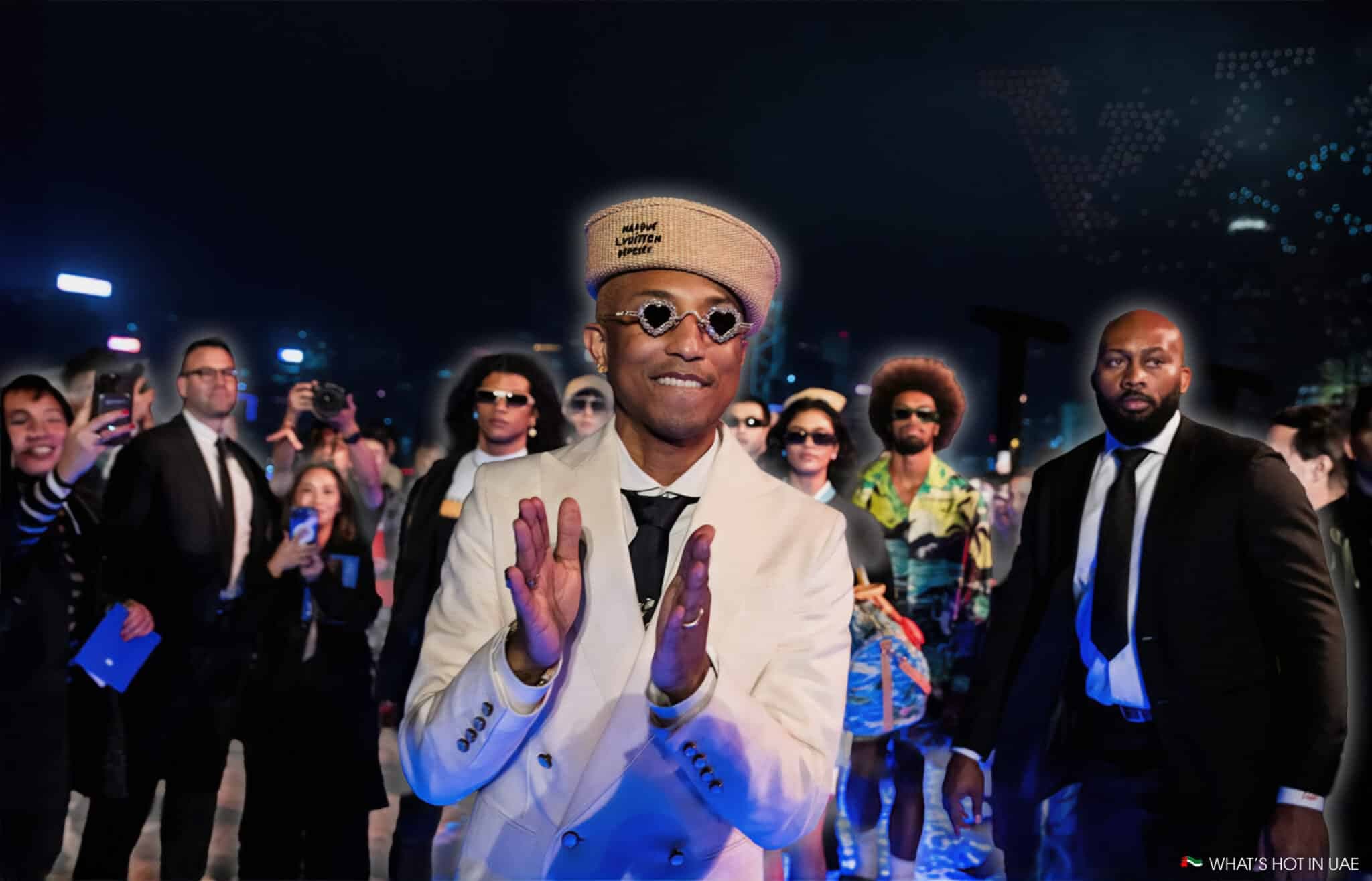 A person in a light-colored suit and beret claps while surrounded by a crowd, some of whom are taking photos, against a nighttime city backdrop with lights.