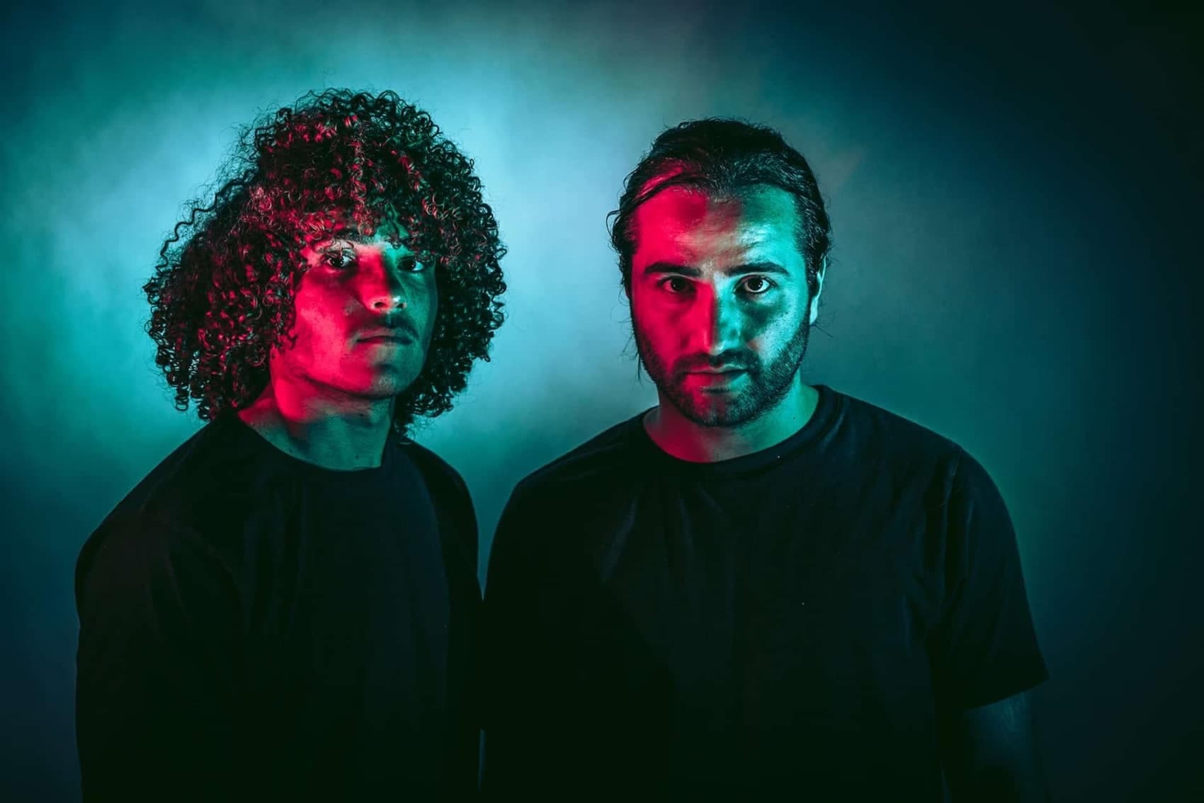 Two people in dark shirts are lit with dramatic red and blue lighting, creating a striking contrast against the dark background.