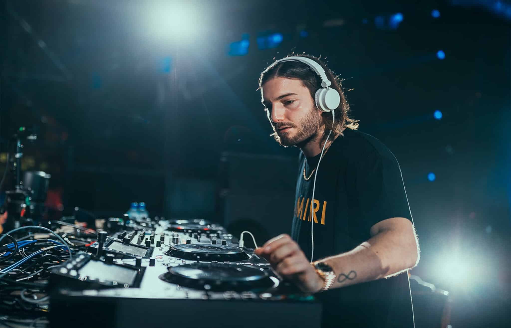 A DJ wearing headphones is focused while mixing music at a DJ booth, illuminated by stage lights.
