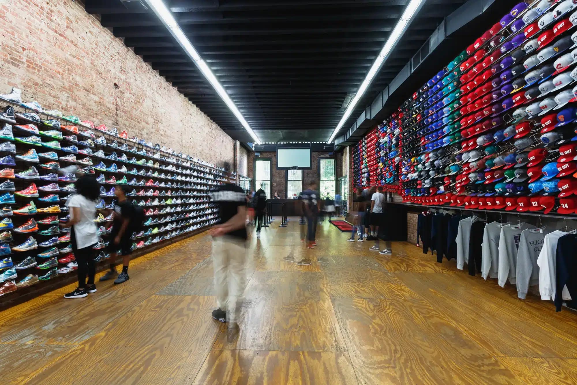 People browsing sneakers and hats on display in a store with brick walls and wooden flooring.