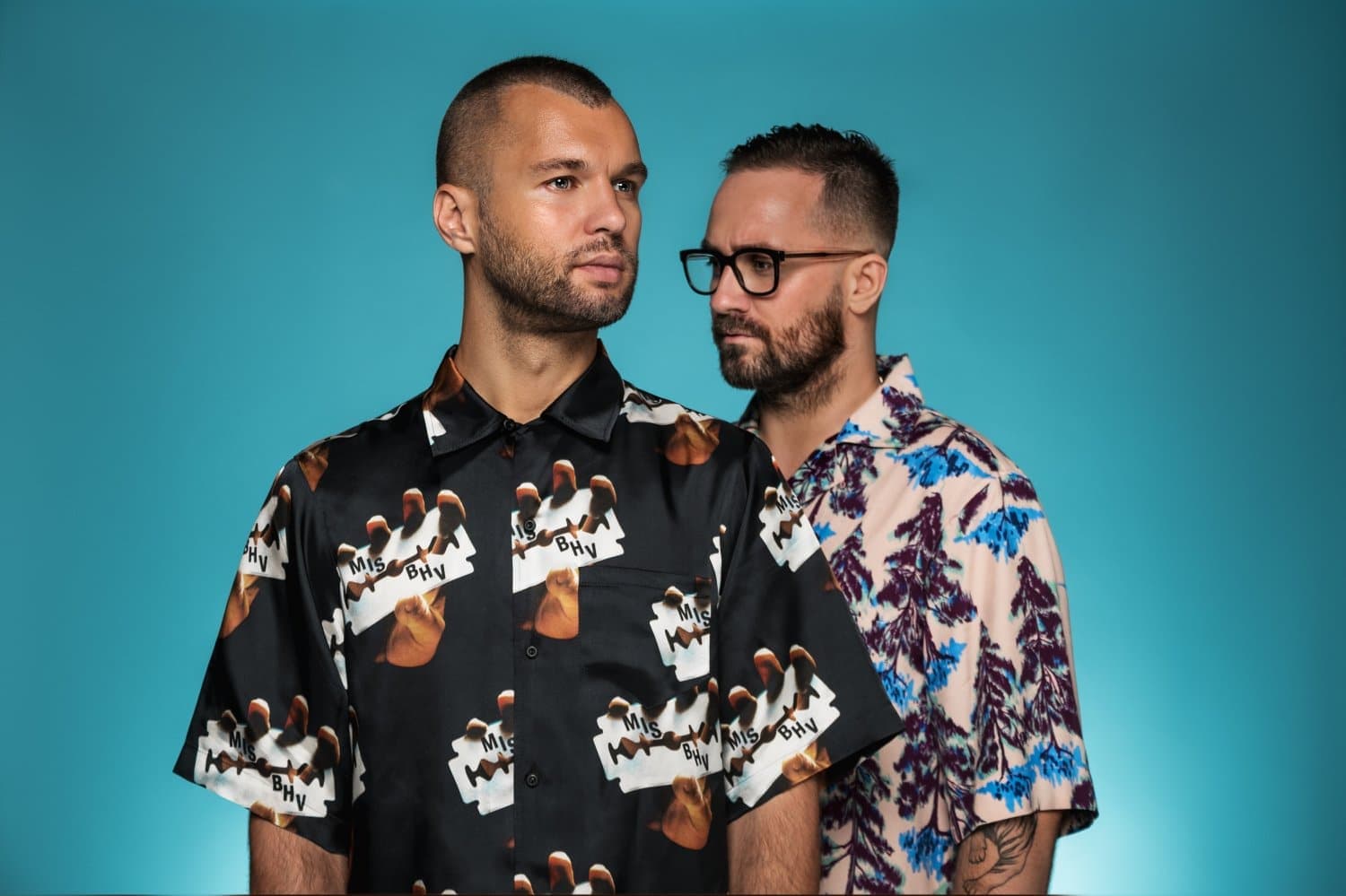 Two men stand against a blue background, one wearing a patterned shirt with photos, the other in a floral shirt.