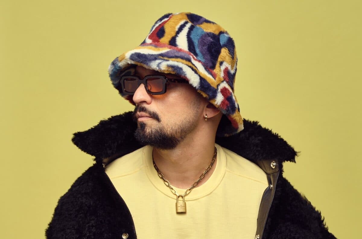 Person wearing a colorful bucket hat, sunglasses, gold necklace, and a black fuzzy jacket against a yellow background.