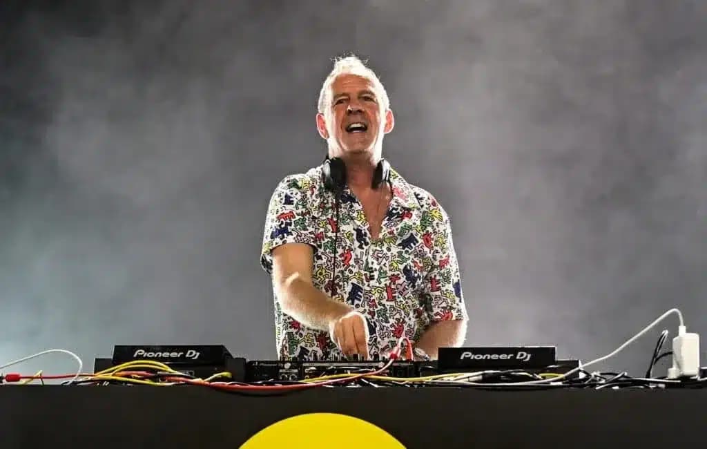 A DJ wearing a colorful shirt performs on stage, surrounded by sound equipment and lighting effects.