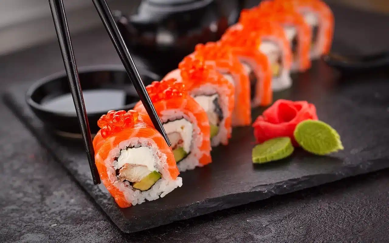 Sushi roll topped with salmon and roe, garnished with a carved tomato rose and served on a black slate plate.